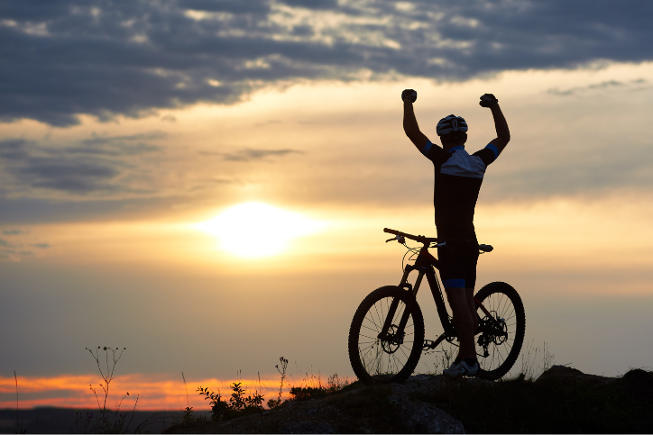 Man in sports clothes and a helmet stands near cycle with his hands up on a rock on top of a mountain